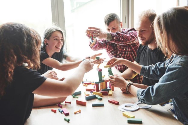 bigstock-A-Group-Of-Creative-Friends-Si-335729296 A group of friends sitting at a wooden table playing a board game.