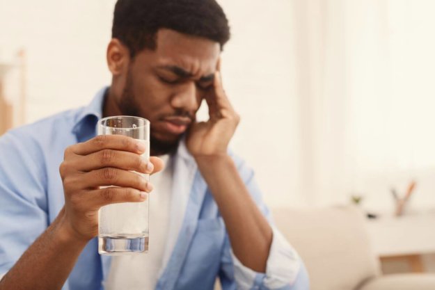 bigstock-Hangover-Upset-African-Americ-293014843 A man holding his head and a glass of water after a night of binge drinking.